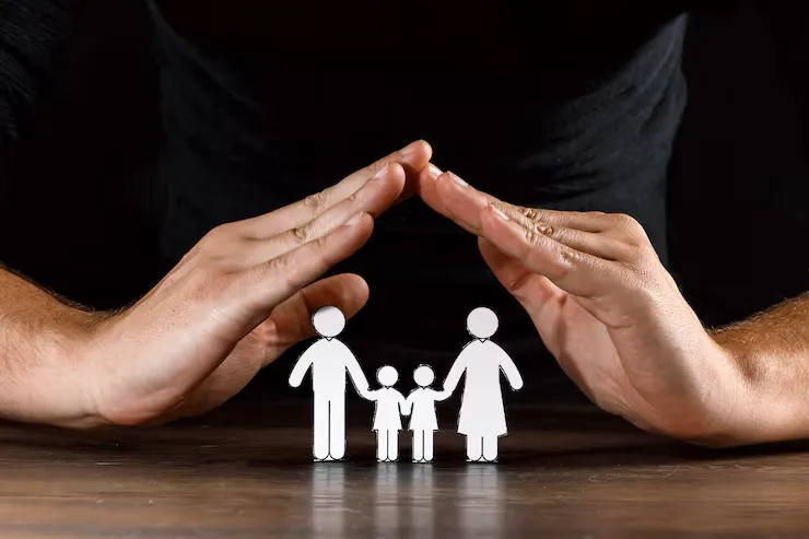 Two hands are cupped protectively over paper cutouts of a family a man, woman, and child, symbolizing care and protection